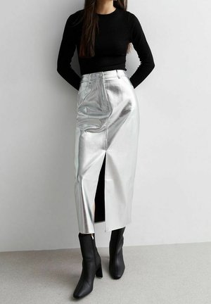 Kvinde iført sort langærmet top, sølvmetalisk midilang nederdel med frontslids og sorte ankelstøvler med hæl, stående mod en grå væg.