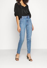 Woman wearing light blue high-waisted skinny jeans, black lace-trimmed camisole, black lace jacket with rolled sleeves, and black heeled sandals.