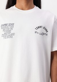 White cotton t-shirt featuring black printed text with city names and brand name. Crew neck design with short sleeves. Smooth texture.