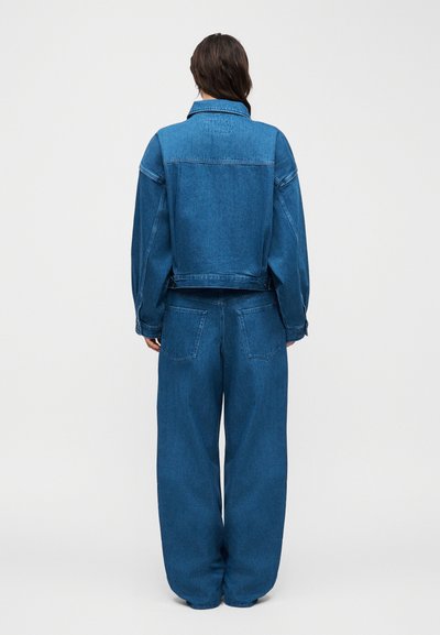 Γυναίκα που στέκεται με το σώμα στραμμένο προς τα πίσω, φορώντας φαρδύ μπλε τζιν μπουφάν και τζιν παντελόνι με φαρδιά πόδια, μπροστά σε απλό ανοιχτό φόντο.