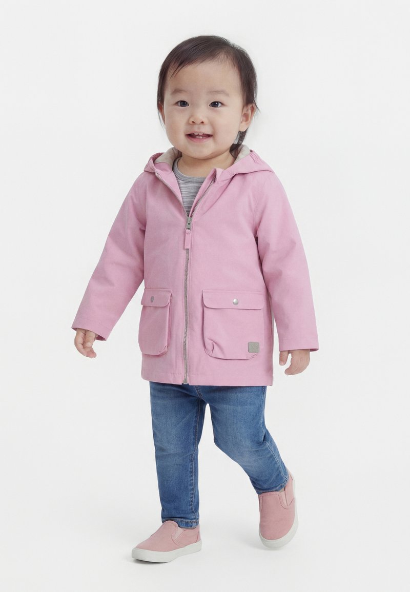Νήπιο που φοράει ροζ μπουφάν με κουκούλα, μπλε τζιν και ροζ παπούτσια slip-on, περπατώντας με ελαφρό χαμόγελο σε λευκό φόντο.