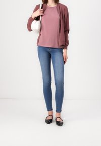 Bordeaux bomberjack over een roze t-shirt, gecombineerd met blauwe skinny jeans en zwarte flats met decoratieve accenten. Witte handtas in de hand.