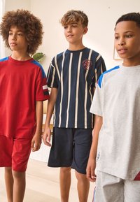 Trois garçons se tiennent ensemble, vêtus de tenues décontractées : une chemise rouge à manches courtes avec des accents bleus, un t-shirt à rayures bleu marine, et une chemise grise avec des détails bleus.