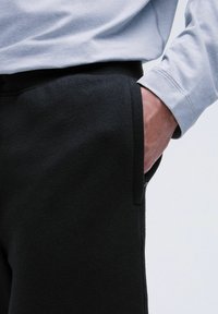 Schwarze Sweatpants mit strukturiertem Oberflächenmaterial, ausgestattet mit Seitentaschen. Kombiniert mit einem hellgrauen Langarmshirt.