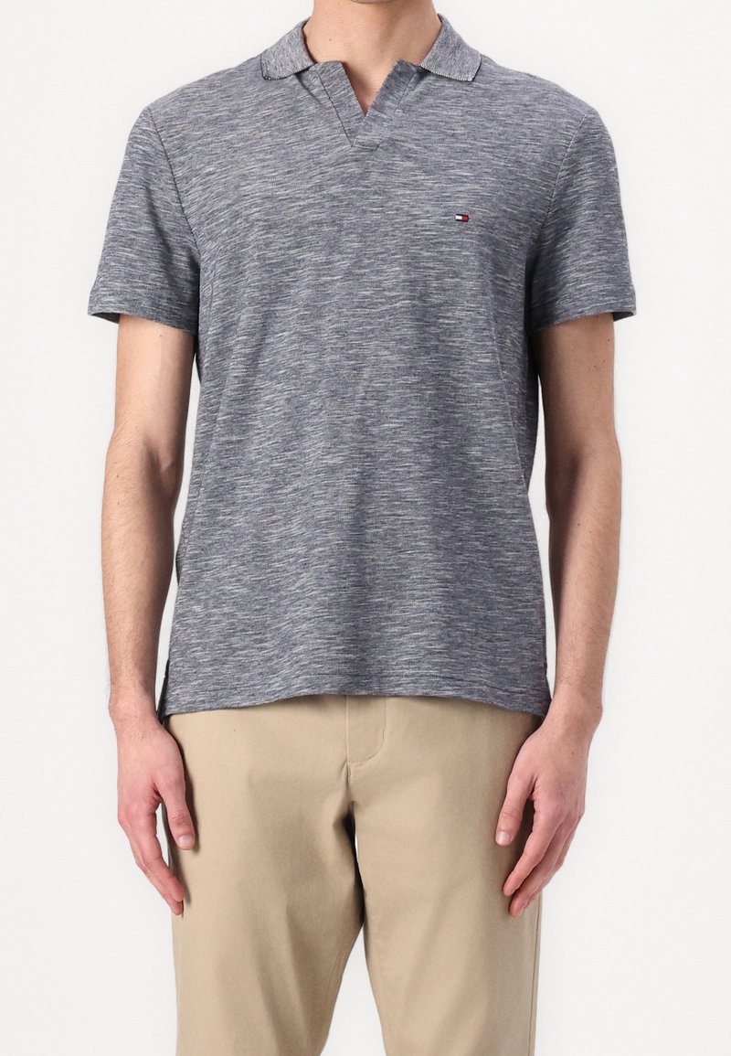 Homme portant un polo gris chiné à manches courtes et un pantalon beige, se tenant devant un fond blanc uni.