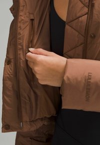Chaqueta acolchada marrón con un bolsillo con cremallera, textura suave y un detalle de logo en la manga. Llevada sobre una prenda negra.