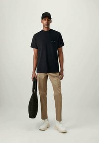 Svart bomull t-skjorte med en liten hvit trykket detalj, beige bukser, hvite sneakers og en teksturert svart veske, båret av en stående mannlig modell.