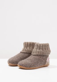 Bruine vilten slippers met een gebreide bovenrand, ronde neus en een gestructureerd oppervlak. Rubberen zool voor grip, met een klein rood merklabel aan de zijkant.