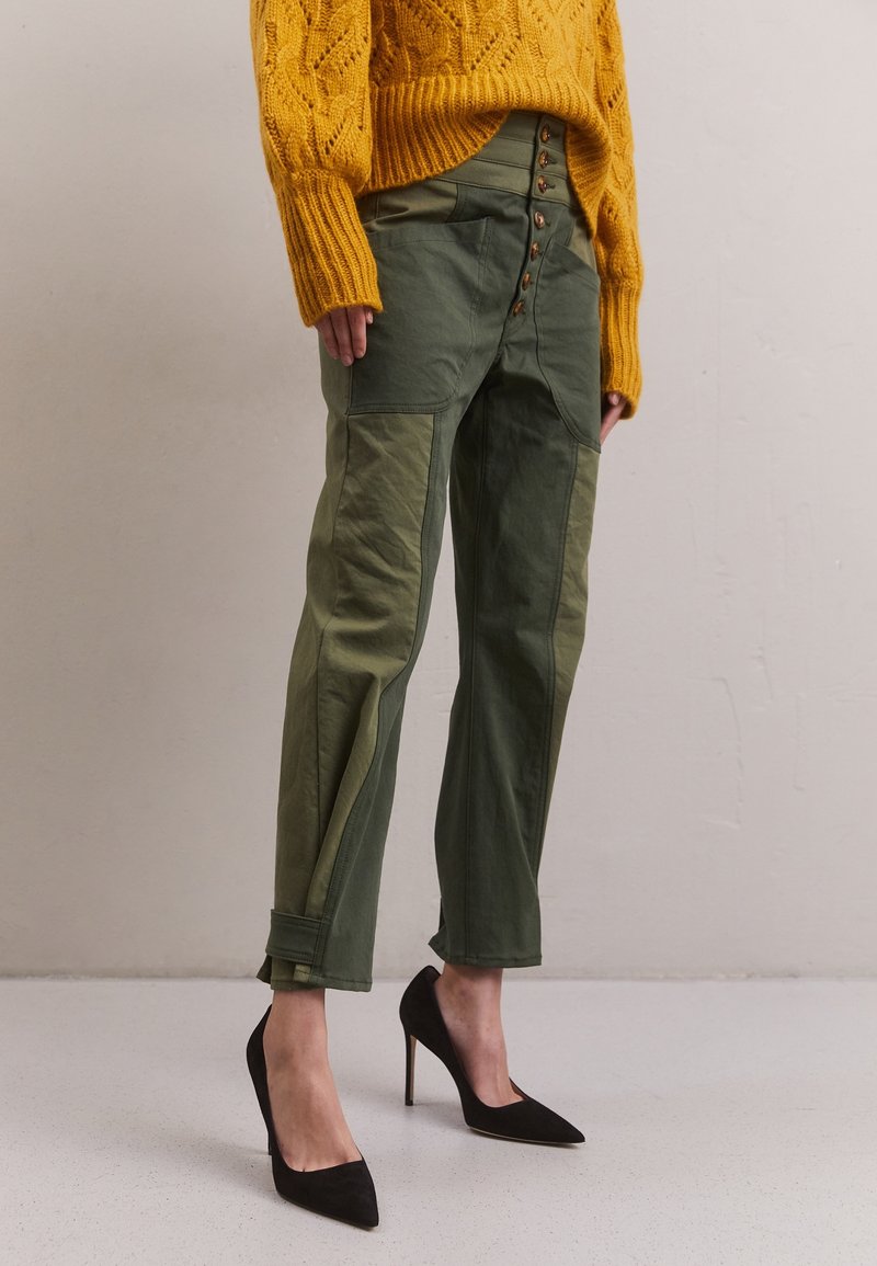 Olijfgroene broek met contrasterende stukken, vijfknops sluiting en opgerolde manchetten, gecombineerd met zwarte puntige hakken en een gele gebreide trui.