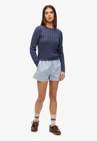 Marineblauwe kabelgebreide trui, lichtblauwe gestreepte shorts, sokken met patroon, bruine schoenen. Casual outfit met gestructureerde materialen en gelaagde stijlen.