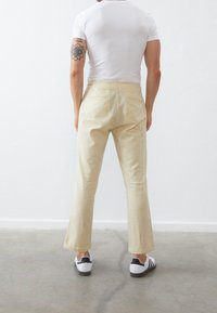 Pantalon beige évasé en tissu léger, avec une texture lisse et deux poches arrière. Porté avec une chemise blanche ajustée et des chaussures.