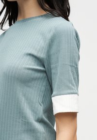 Γυναίκα που φοράει γαλάζιο τοπ με ρίγες και λευκά μανσέτα στα μισά μανίκια, εμφανιζόμενη από τον λαιμό μέχρι το μέσο του κορμού.