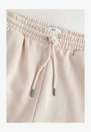 Hellbeige Jogginghose mit Kordelzug am Bund. Weiche Textur, elastischer Bund mit gerafften Details und silberfarbene Metallenden an der Kordel.