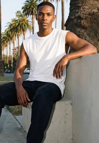 Mladi moški, oblečen v belo majico brez rokavov in črne hlače, sedi na betonski ograji, v ozadju pa so palme in cesta.