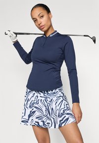 Maglietta da golf a maniche lunghe color navy con colletto a zip, abbinata a una gonna bianca con motivi astratti blu e pieghe. Mazza da golf tenuta in mano.