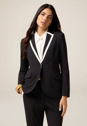 Schwarzes Blazer mit weißen Akzenten, taillierte Form, zwei Taschen, kombiniert mit einem weißen Hemd. Glattes Material, strukturierte Silhouette und minimale Details.