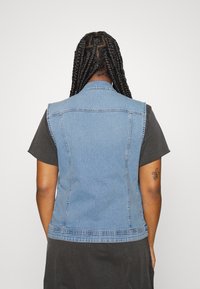 Denim brezrokavnik z svetlo modrim umivanjem, brez rokavov, s špičastim ovratnikom in kontrastnimi šivi, oblečen čez črno majico.