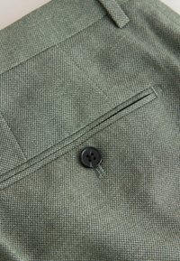 Nærbillede af grøn struktureret stof med en sort firehuls knap og en knaphul på en skræddersyet lomme på et tøjstykke.
