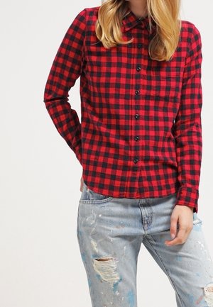 Femme portant une chemise à carreaux rouge et noire boutonnée et un jean bleu clair usé avec des éclaboussures de peinture, debout avec une main dans la poche.