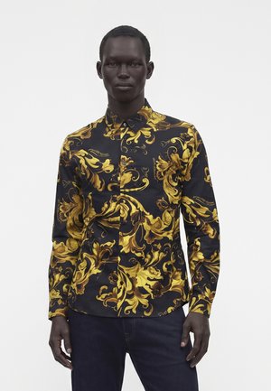 Man draagt een zwart overhemd met knopen met een sierlijk geel en goud bloemrijk barokpatroon, gecombineerd met donkerblauwe broek.