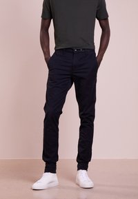 Pantaloni blu navy con una vestibilità slim, caratterizzati da tasche laterali e una texture liscia, abbinati a sneakers bianche e a una t-shirt aderente di colore scuro.