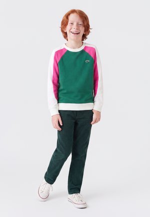 Enfant roux souriant portant un sweat-shirt vert et rose, un pantalon vert foncé et des baskets blanches sur un fond clair uni.