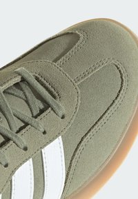 Zeleni sued cipelica s bijelim kožnim prugama, s округlim vrhom, vezanjem na vezice i gumiranim potplatom. Glatka tekstura s vidljivim šavovima.