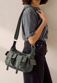 Zielona nylonowa torba na ramię z wieloma przednimi kieszeniami i regulowanym paskiem, charakteryzująca się gładką fakturą i przeszyciami.