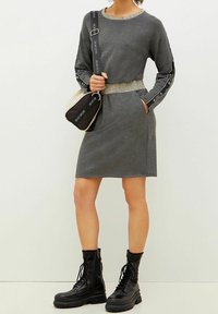 Robe grise à manches longues avec une ceinture scintillante, assortie à des bottes noires à lacets épais et à un sac bicolore avec des accents de logo.