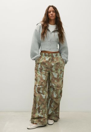 Femme aux longs cheveux bouclés, portant un sweat à capuche court gris avec zip, un pantalon camouflage ample et des chaussures plates blanches, debout devant un fond uni.