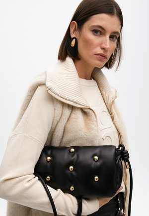 Borsa in pelle nera con borchie dorate, texture morbida, manico annodato, tenuta da una persona in un outfit beige con giacca foderata in pelliccia.