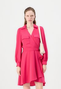 Abito avvolgente rosa brillante con maniche lunghe, due tasche sul petto, vita legata e una gonna fluida. Il modello tiene una piccola borsa beige.