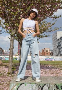 Jeans de perna larga azul claro com um joelho rasgado, combinados com um top cropped lilás com nó na frente e ténis brancos. Chapéu de balde rosa como um acessório.