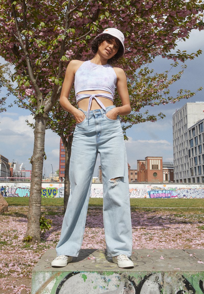 Jeans de perna larga azul claro com um joelho rasgado, combinados com um top cropped lilás com nó na frente e ténis brancos. Chapéu de balde rosa como um acessório.