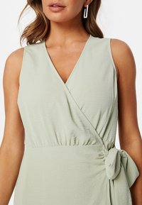 Svetle zelené zavinovací šaty s hlubokým výstřihem do V, s nařaseným pasem a zavázaným detailem na boku. Měkká texturovaná látka s volným designem.