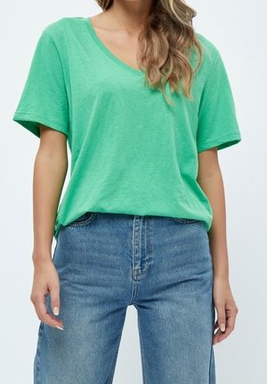 Woman wearing a loose green V-neck T-shirt tucked into high-waisted blue jeans, standing against a plain background.