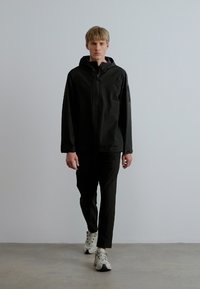 Fekete könnyű dzseki kapucnival, elöl cipzárral és texturált anyagból, fekete nadrággal és fehér tornacipőkkel párosítva. Sima, modern dizájn.