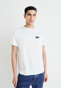 Biela krátkouramenná tričko s okrúhlym výstrihom a malým modrým obdĺžnikovým logom na ľavej hrudi. Ľahký jersey materiál.