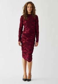 Burgunderfarbenes Langarmkleid mit floraler Stickerei, gerafftem Design und taillierter Silhouette, kombiniert mit schwarzen High Heels.