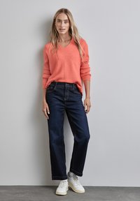 Korbmuster-Strickpullover mit vertikalen Rippungen, kombiniert mit dunkelblauen weit geschnittenen Jeans und weißen Turnschuhen. Der Hintergrund ist neutral grau.