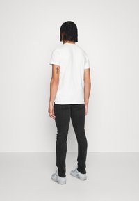 Camiseta blanca de manga corta, jeans negros ajustados y zapatillas gris claro. El sujeto está mostrado de espaldas, con un tatuaje visible en el brazo izquierdo.