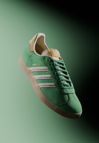 Sneaker verde in suede con strisce bianche, caratterizzata da un accento testurizzato sul tallone e una linguetta di colore chiaro. Lacci piatti e suola in gomma.