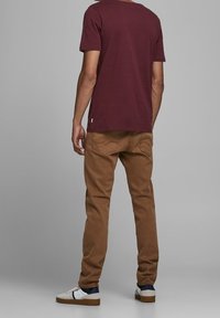 Un homme de dos portant un t-shirt bordeaux, un pantalon marron et des baskets blanches avec des rayures bleu marine et rouge.