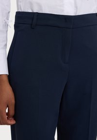 Pantalon taillé navy avec une texture lisse, doté d'un devant plat, de passants de ceinture et d'une coupe ajustée. La main repose légèrement sur le tissu.