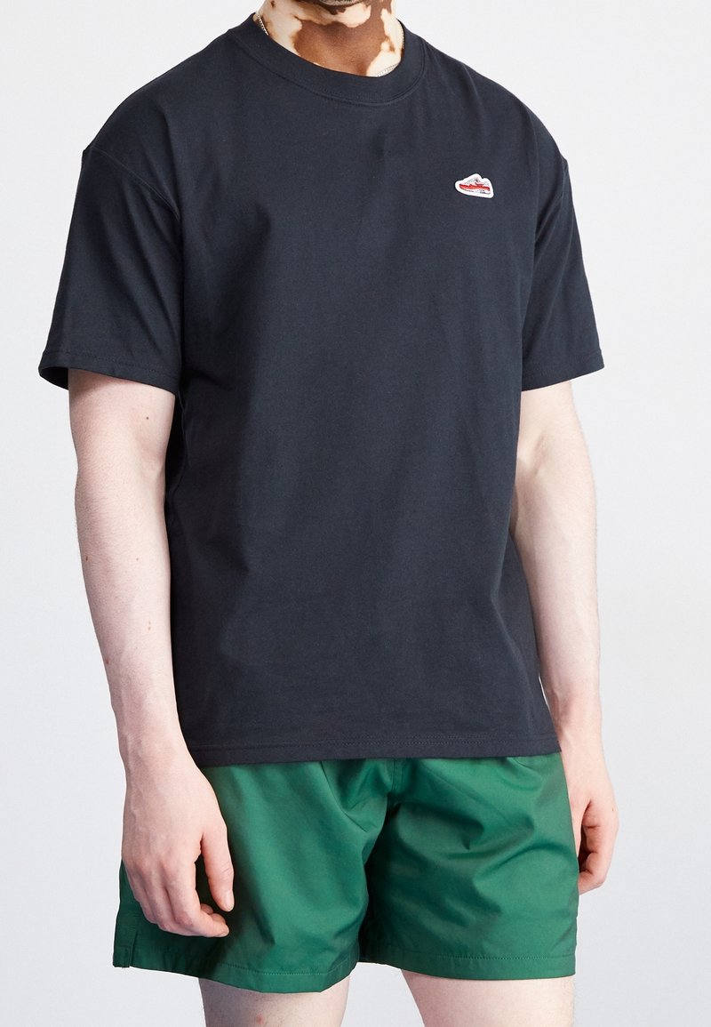 Svart bomullströja med kort ärm och rundad halsringning, med en liten röd och vit logga på bröstet. Matchas med gröna shorts.