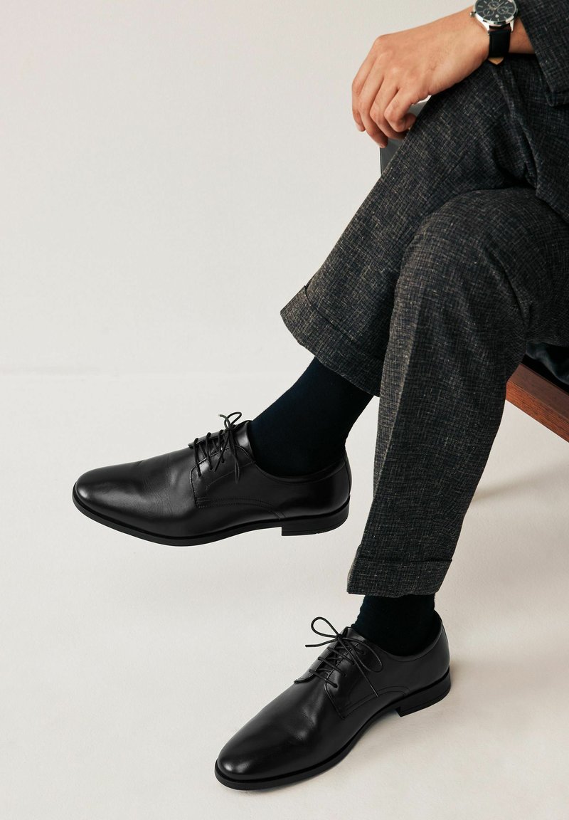 Zapatos de vestir de cuero negro con punta redonda y diseño de cordones, combinados con calcetines oscuros y pantalones de textura gris. Estilo minimalista con acabado suave.