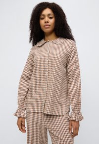 Καρό πουκάμισο σε καφέ και άσπρο χρώμα με γιακά, μακριά μανίκια και μανσέτες με βολάν. Ελαφρύ ύφασμα, κλείνει με κουμπιά μπροστά και έχει άνετη εφαρμογή.