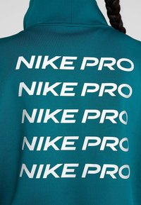 Veste de sport bleu sarcelle avec texte blanc répété "NIKE PRO" dans le dos, portée par une personne avec une coiffure longue tressée.