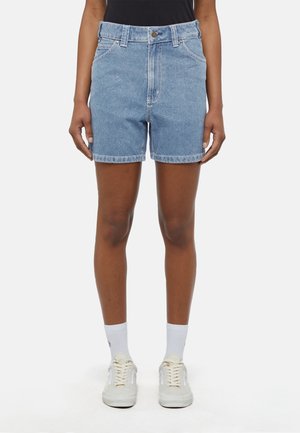 Persona con shorts vaqueros azul medio, calcetines blancos hasta la mitad de la pierna y zapatillas blancas, de pie frente a un fondo liso.