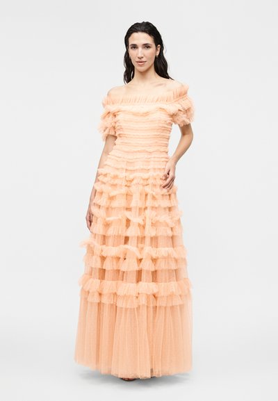 Naine kannab virsikuvärvilist, õlgadeta, astmelist tuulekleiti volangide detailidega ja seisab lihtsa heleda tausta ees.